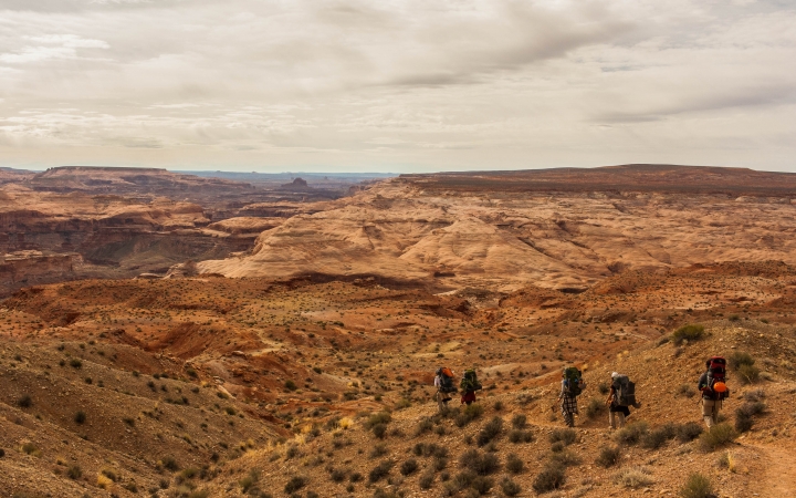Five students wearing backpacks hike along a trail is a vast desert landscape.